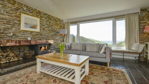 A cozy living room with a stone fireplace, a sofa, a coffee table, and a large window offering a view of the hills. - Ayrmer House (Kingsbridge)