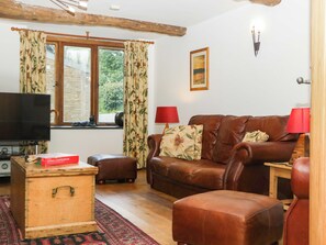 A living room with a brown leather sofa, a wooden chest, a television, and a window with curtains. - The Hay Barn (Totnes)