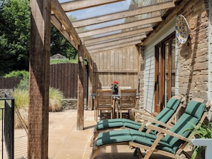 A wooden pergola with a table and chairs, a stone wall, and a green cushioned lounge area.