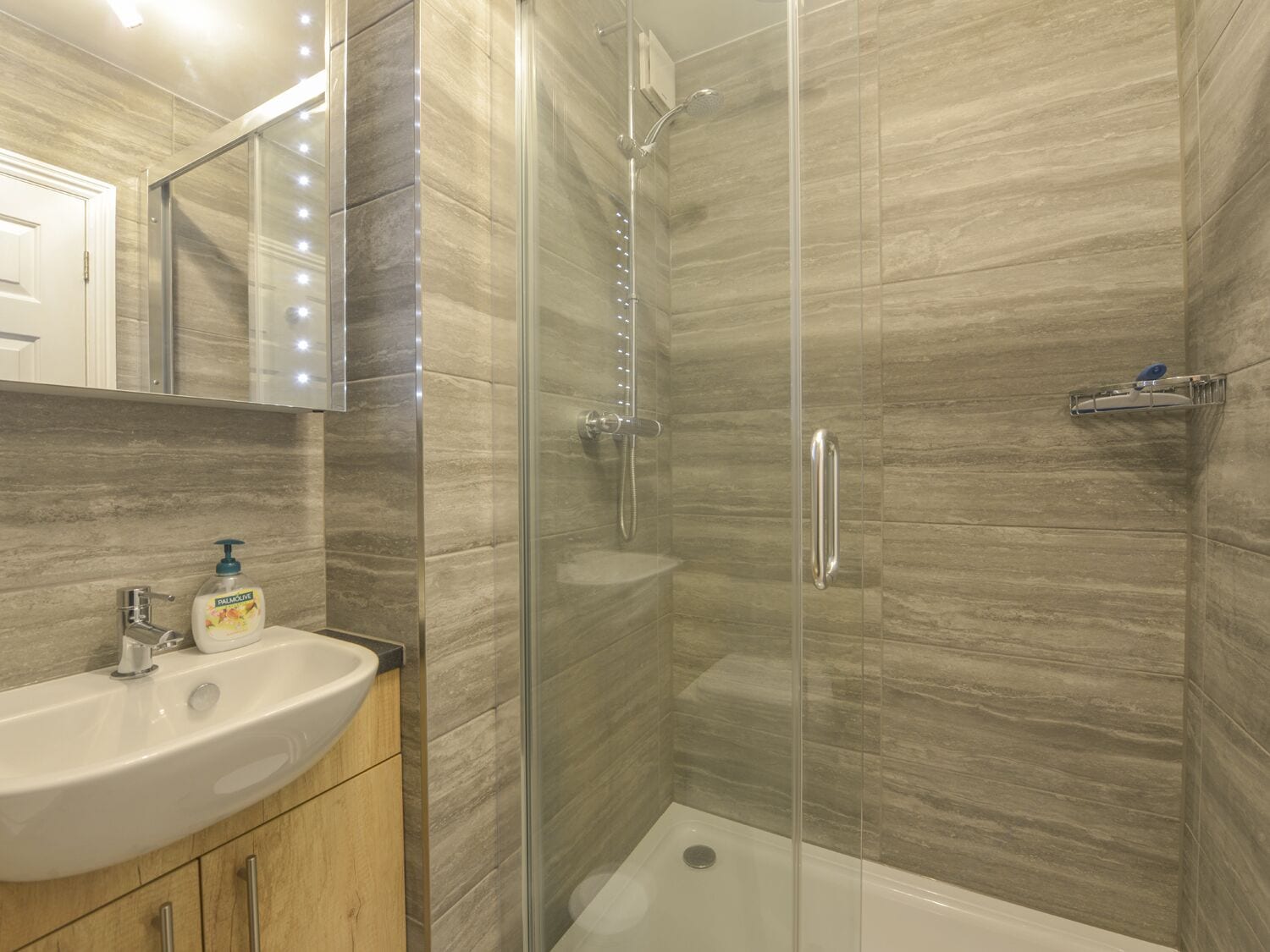 Une salle de bain moderne dotée d’une douche à parois de verre, d’un meuble-lavabo en bois et d’un miroir avec éclairage intégré.