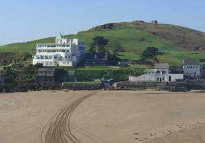 A coastal building with multiple floors, situated on a sandy beach with a hill in the background.