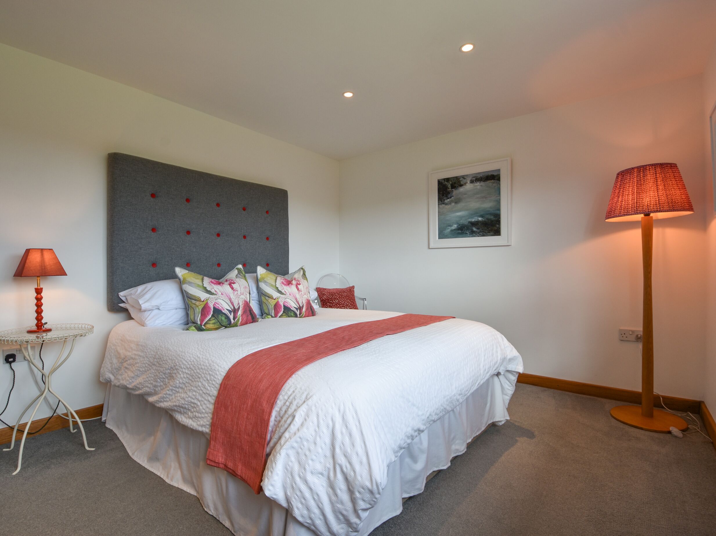 A neatly arranged bedroom with a large bed, a grey headboard, a red lamp, and a framed picture on the wall.