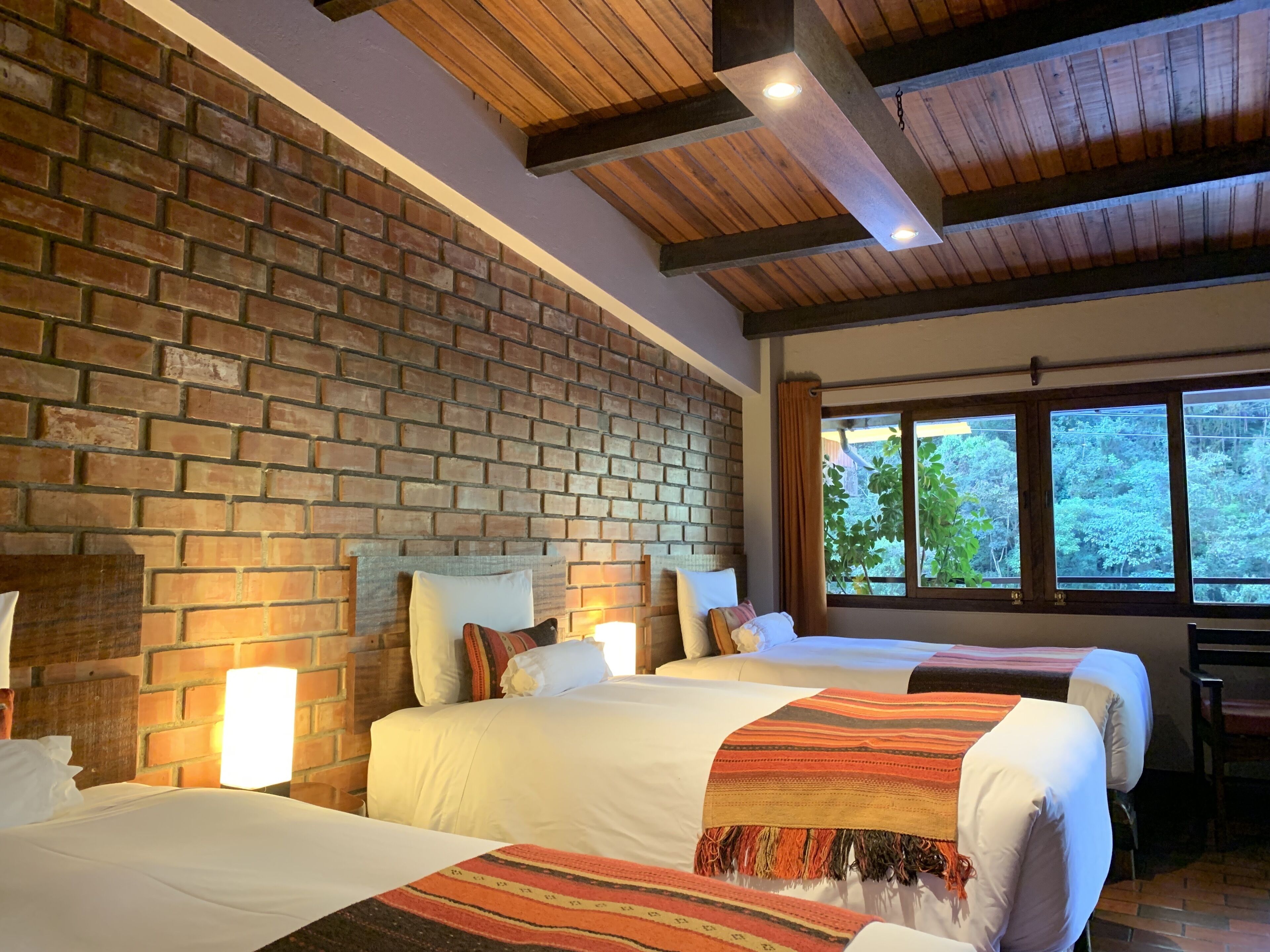 A hotel room with a brick wall, wooden ceiling beams, two beds with white linens, and a large window with a view of trees.
