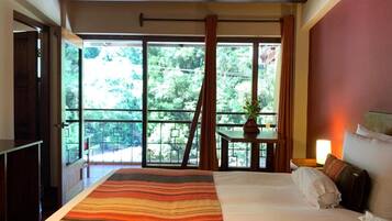 A hotel room with a large bed, a view of greenery through the window, and a small table with a plant.