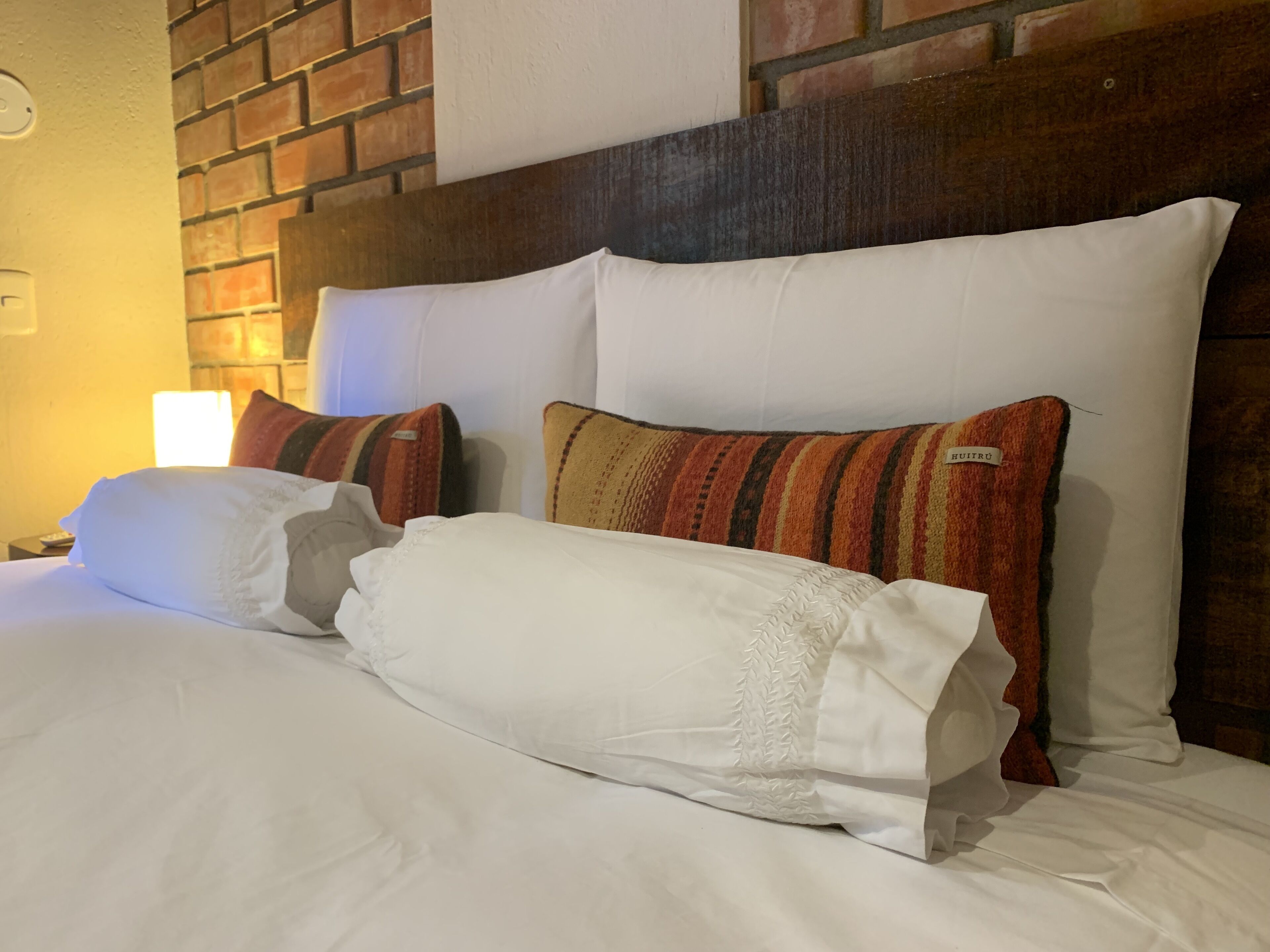 Un lit bien fait, avec du linge de lit blanc et des oreillers à rayures, une tête de lit en bois et un mur en briques en arrière-plan.