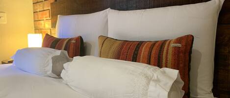 A neatly made bed with white linens and striped pillows, a wooden headboard, and a brick wall behind.