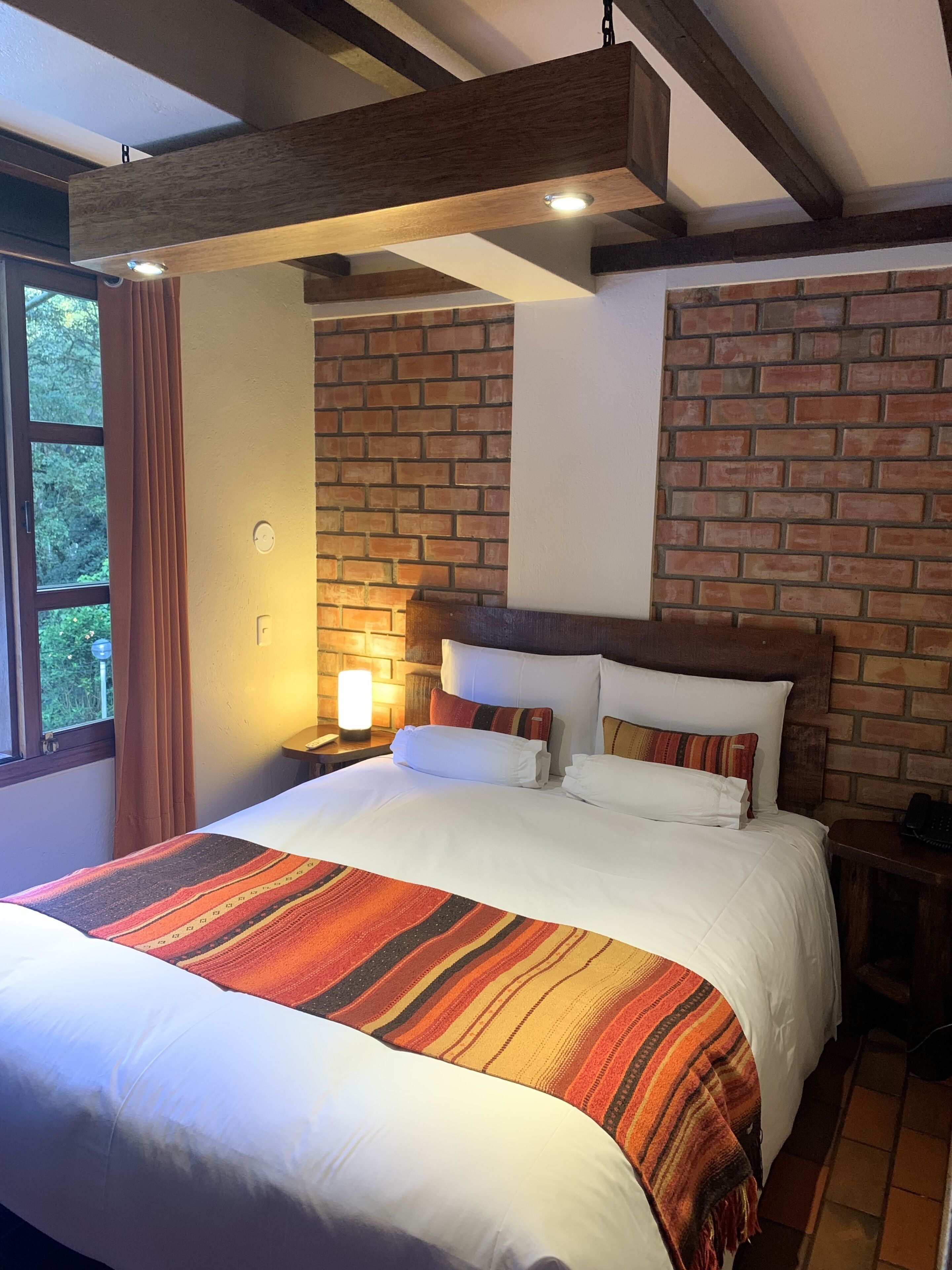 A neatly made bed with a striped blanket, a brick-patterned wall, and a wooden ceiling beam.