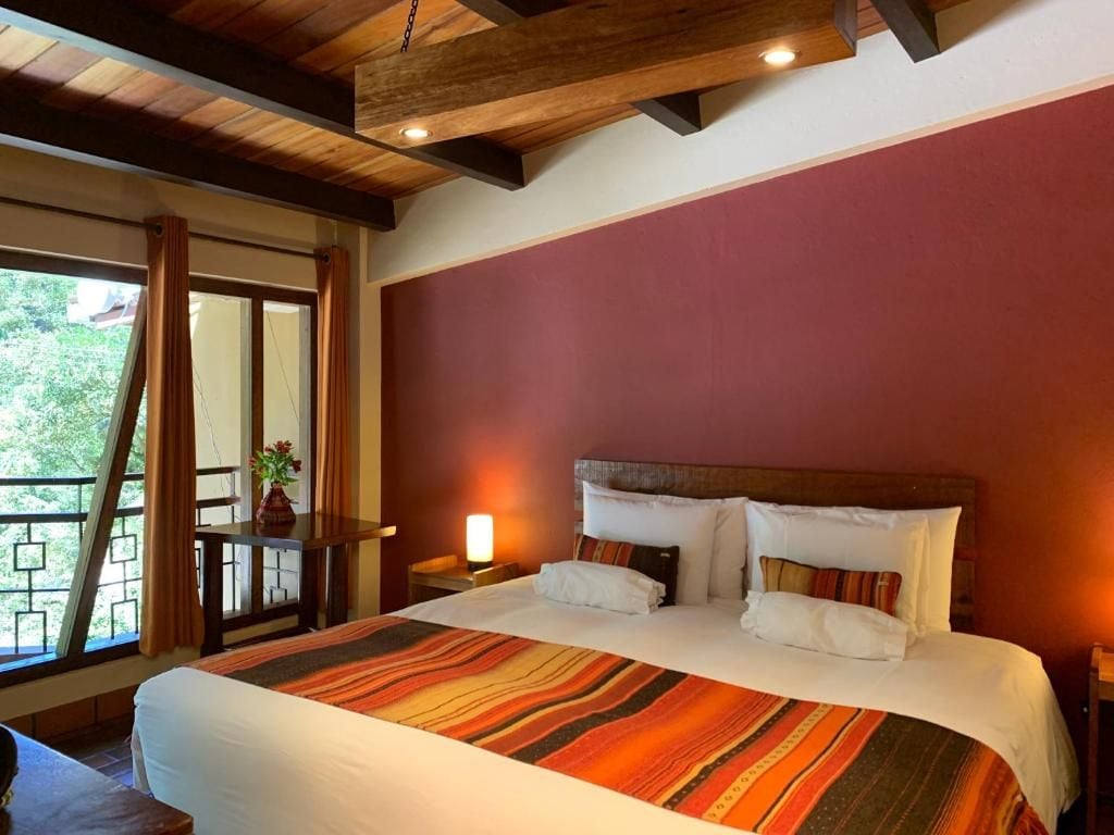 Ein Hotelzimmer mit einem großen Bett, Holzbalkendecke, Blick durch ein Fenster und einem Nachttisch mit einer Lampe.