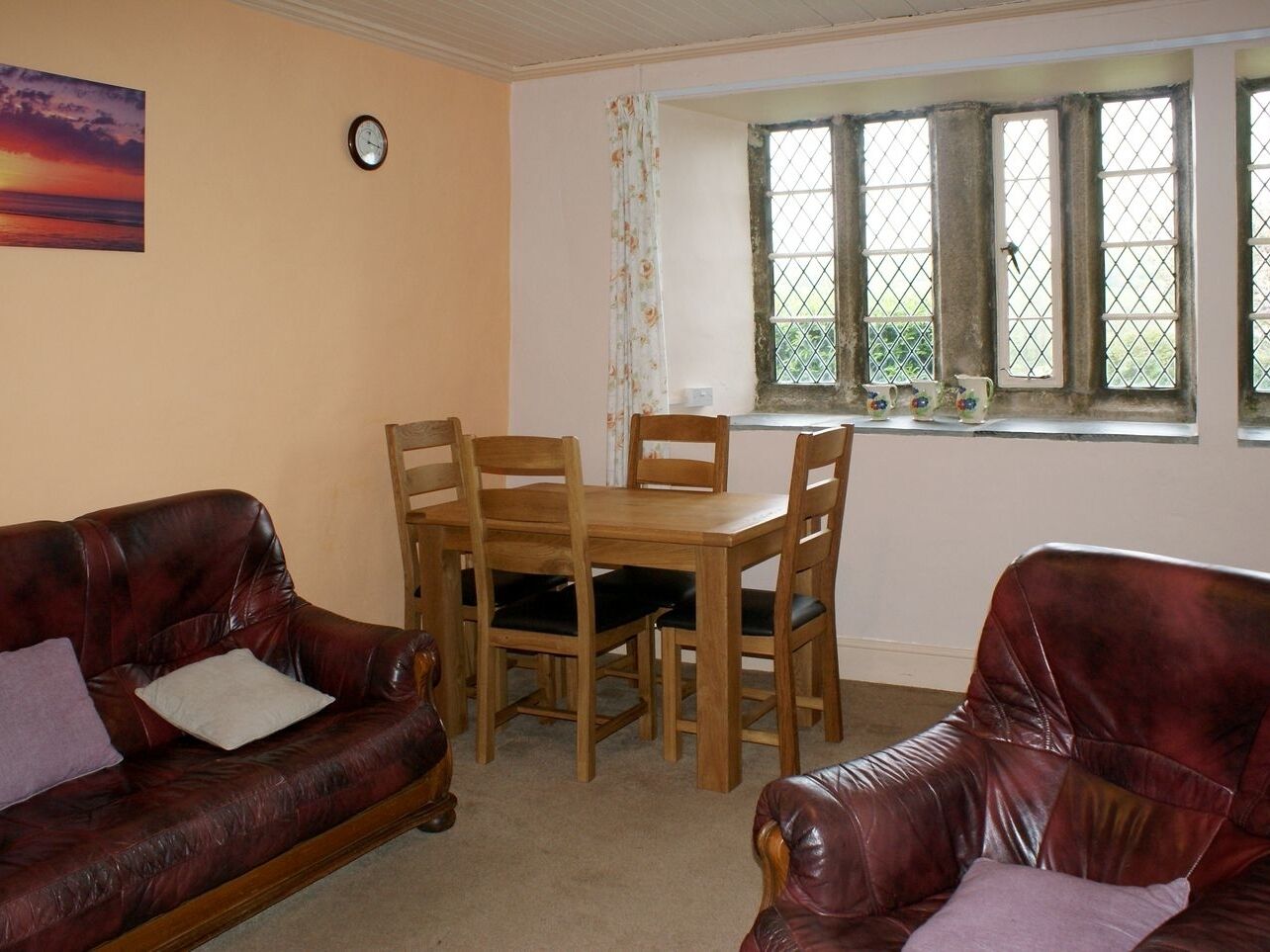 A living room with a wooden dining table and four chairs, a red leather sofa, and a window with a view of greenery.