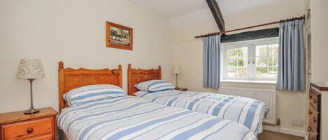 Quarto com duas camas, cabeceiras de madeira, mesas de cabeceira com luminárias, uma janela com cortinas e um radiador.