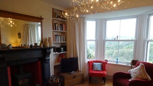 Ein gemütliches Wohnzimmer mit Kamin, Bücherregalen, einem Fernseher und Blick ins Grüne durch große Fenster.
