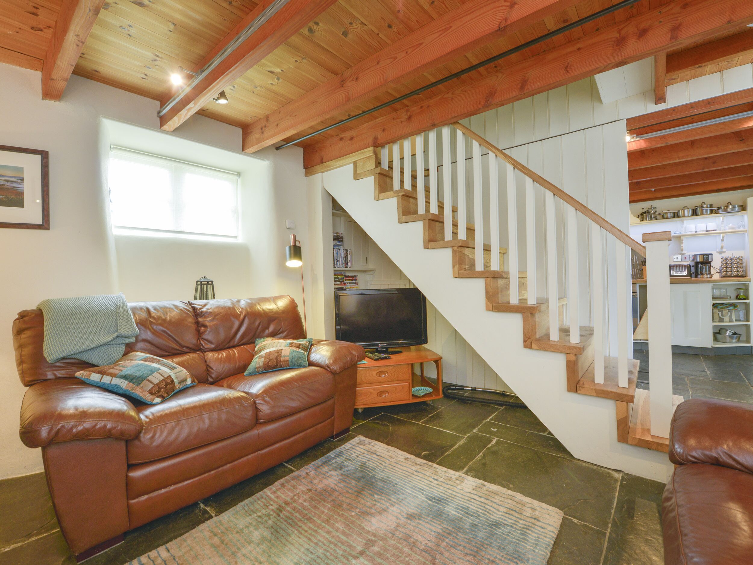 A cozy living room with a brown leather sofa, a television, and a staircase leading to a kitchen area.