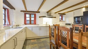A kitchen with white cabinets, a stone tile floor, wooden beams, and a dining area with wooden chairs. - Samuels Cottage (Looe)