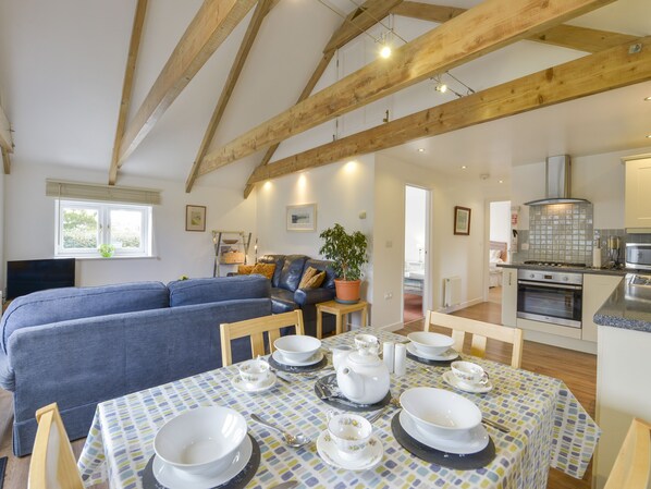 A cozy living room with a blue sofa, a dining table set with white dishes, and a kitchen area with modern appliances. - Hideaway Barn (St Austell)
