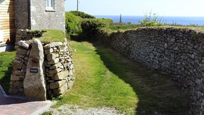 A stone wall leading to a stone cottage with a small window, a stone plaque labeled "THE STUDIO," and a grassy path. - The Studio (Helston)