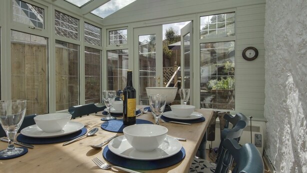 A conservatory with a wooden table set for dining, large windows, and a view of the garden. - 11 The Green (Budleigh Salterton)