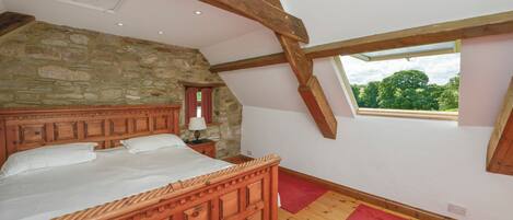 A bedroom with a stone wall, wooden ceiling beams, a large bed, a red rug, and a window with a view of greenery.