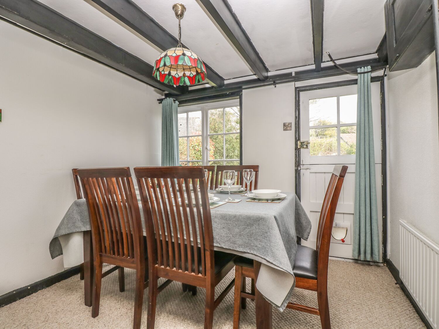 A dining area with a table set for four, wooden chairs, a chandelier, and a view through a window with curtains.