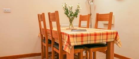 A dining area with a table set for four, wooden chairs, a plaid tablecloth, a vase with flowers, and a wall clock.