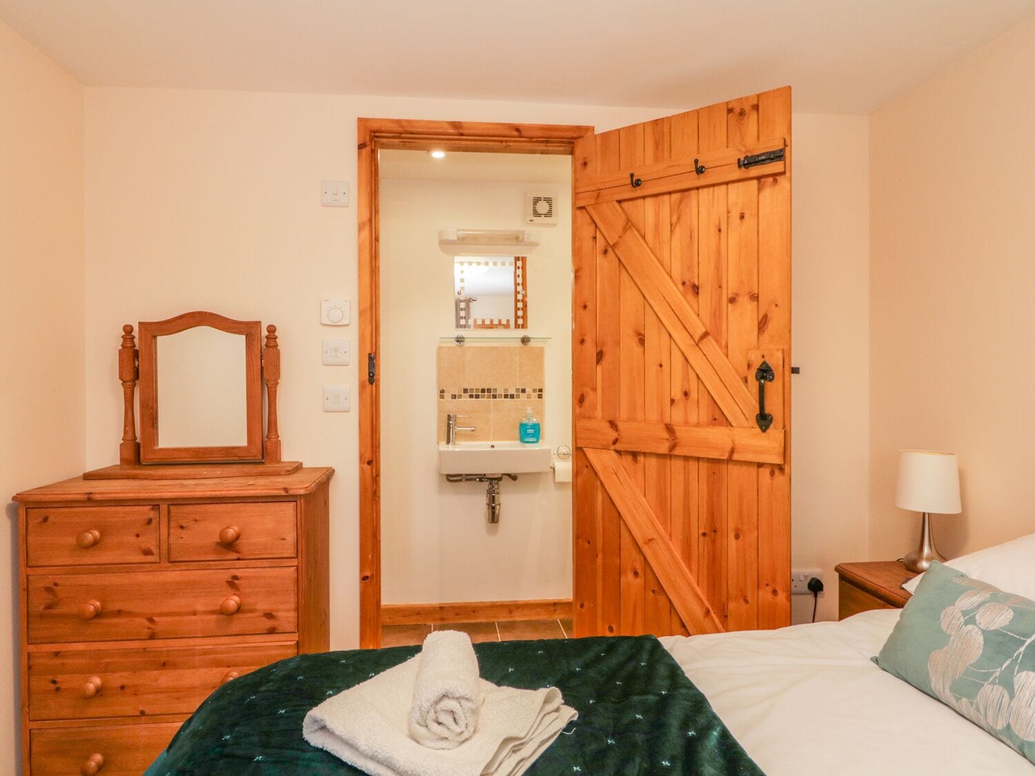 A bedroom with a wooden dresser, a bed with a green blanket, a wooden door, and a nightstand with a lamp.