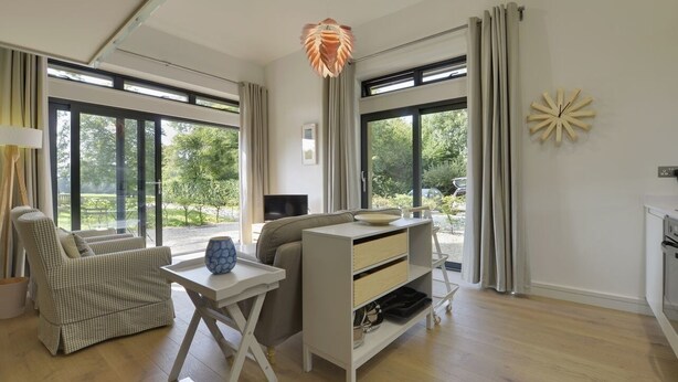 A modern living room with a sofa, a desk, a lamp, and a view of greenery outside. - Grooms Cottage (Exeter)