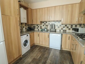 A compact kitchen with wooden cabinets, a washing machine, a dishwasher, a microwave, and a gas stove. - Halford Hill (Caernarfon)