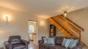 Un salon avec un canapé en cuir marron, un fauteuil assorti, un escalier en bois et une cuisine visible à travers une porte ouverte.