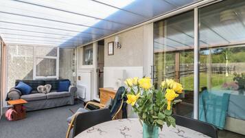 A covered patio area with a dining table, chairs, a sofa, and a vase of yellow flowers.