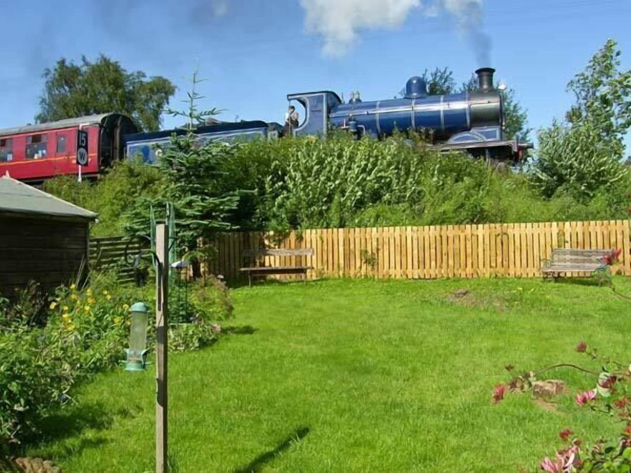 Un jardín con cerca de madera, un banco y un tren rojo al fondo.
