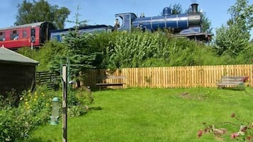 Un jardín con cerca de madera, un banco y un tren rojo al fondo.