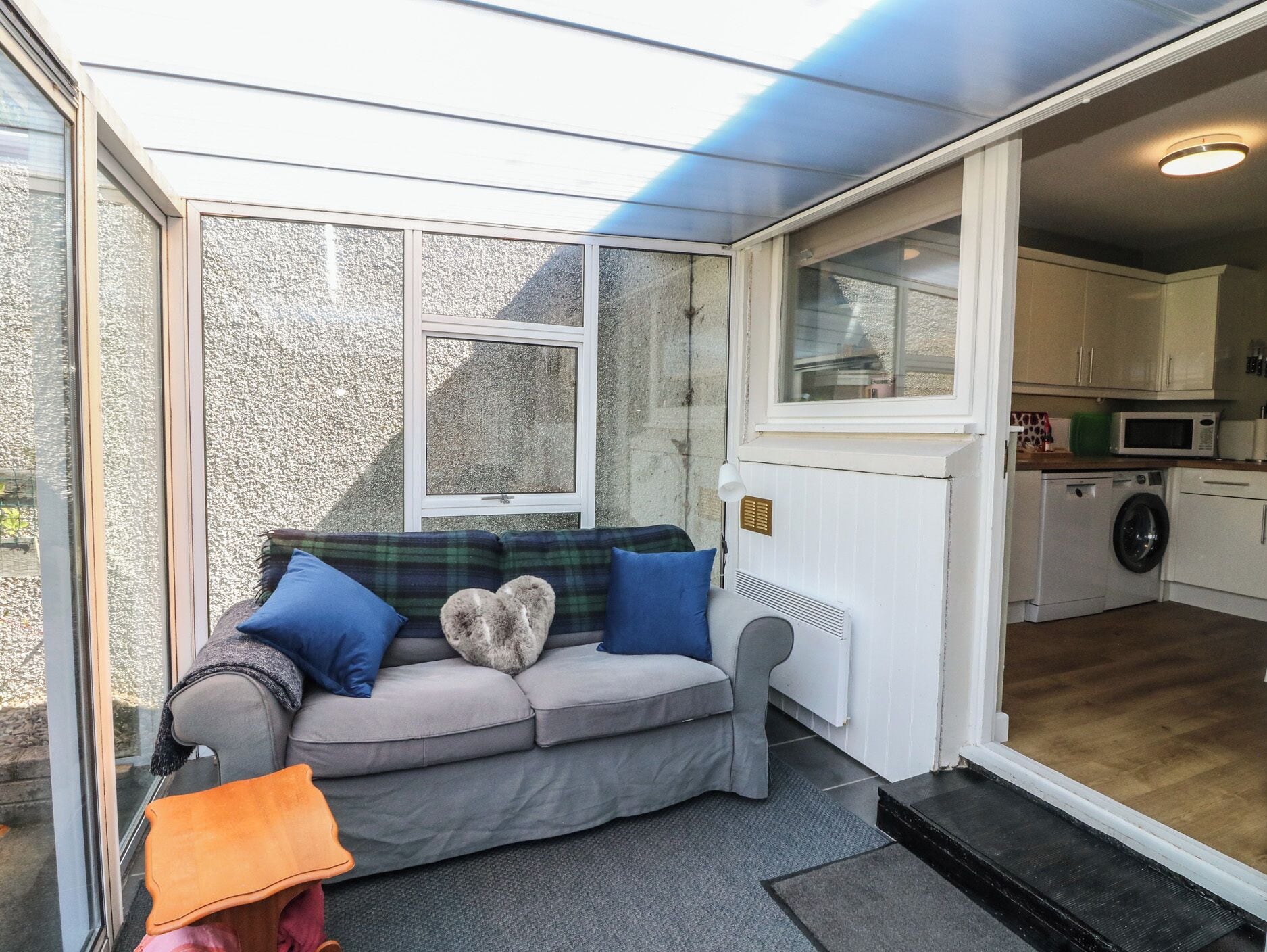 Un cuarto acogedor con un sofá gris, un sillón naranja pequeño, una ventana y una cocina visible a través de una puerta abierta.