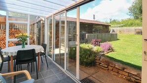 Een overdekt terras met een tafel en stoelen, een stenen muur, en een tuin met bloemen en een houten omheining.