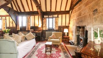 Une chambre traditionnelle avec des poutres apparentes, une cheminée, un grand tapis et un lit agrémenté de nombreux coussins.