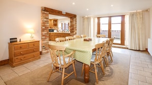 A dining room with a large table, wooden chairs, a brick wall, and a view through a sliding door. - Hadleigh Farm Barn (King's Lynn)