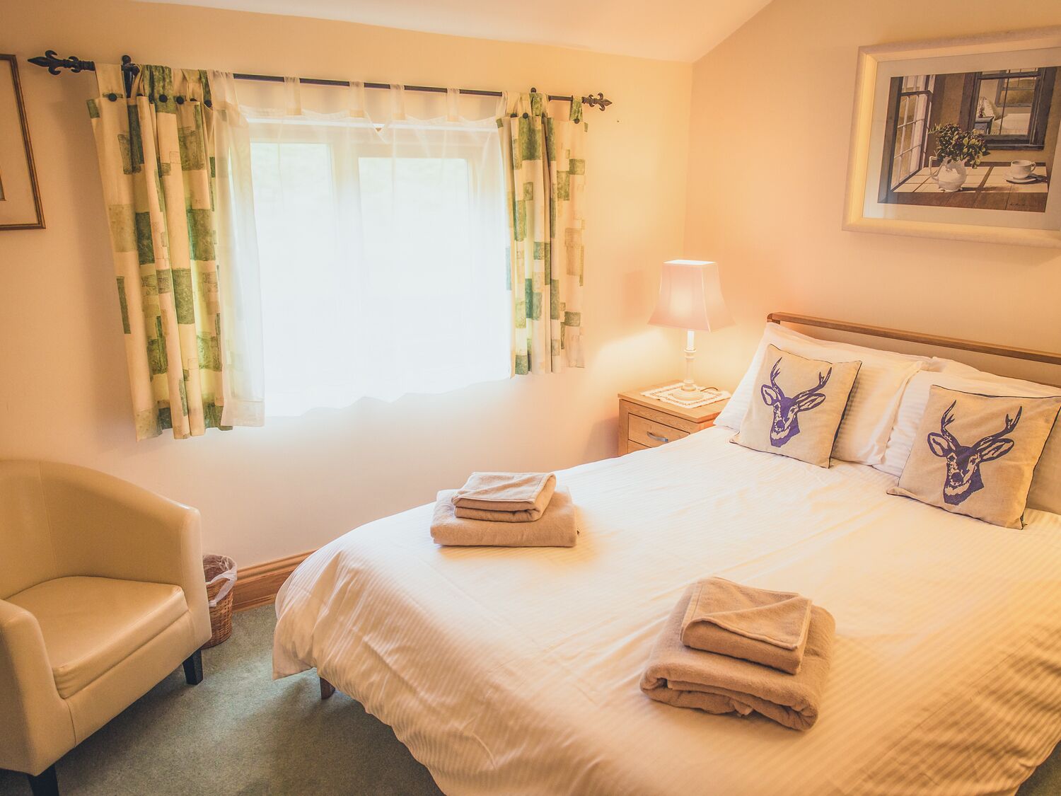 A neatly made bed with pillows, a bedside lamp, and a chair in a room with a window and curtains.