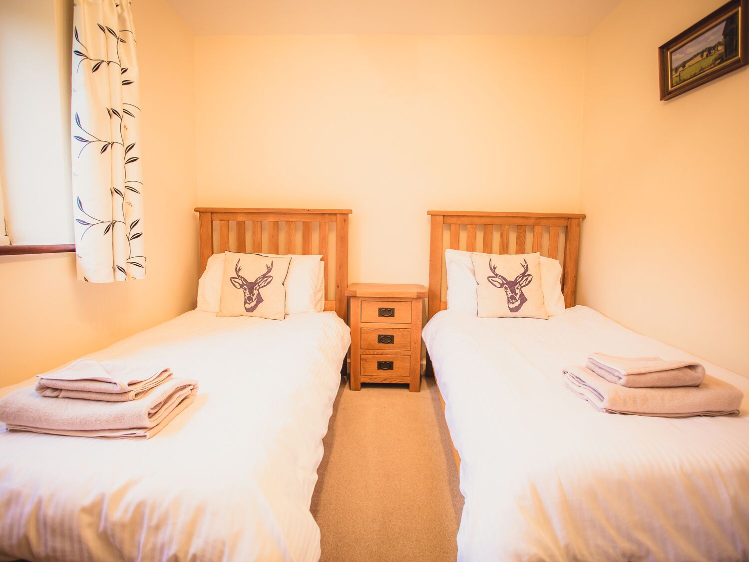 A double room with two beds, each with a pillow featuring a deer head, a wooden nightstand with drawers, and a window with a curtain.