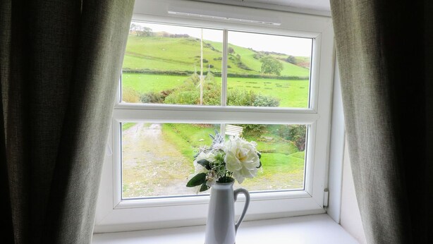 A window with a white vase containing flowers, curtains, and a view of a green landscape. - Penglanowen Fawr (Aberystwyth)