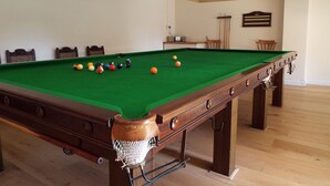 Una mesa de billar con superficie de fieltro verde, rodeada de sillas de madera y una mesita con un radio.