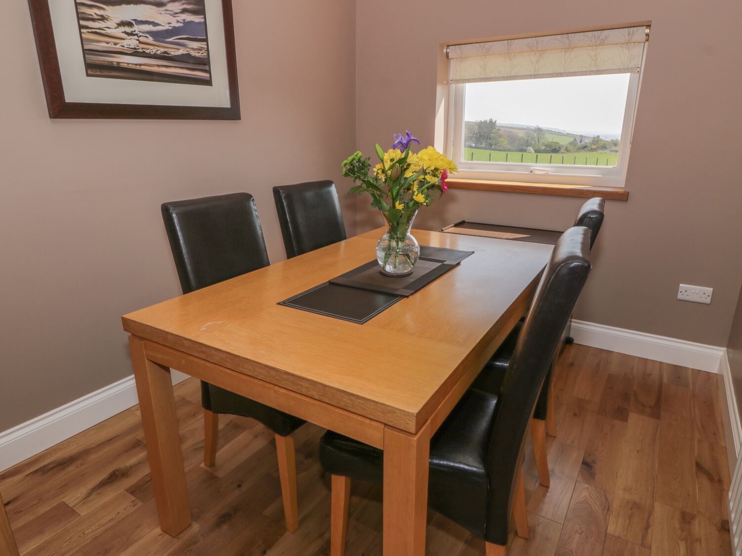 Uma área de refeições com uma mesa de madeira, cadeiras pretas, um vaso com flores, um quadro na parede e uma janela com vista para o campo.