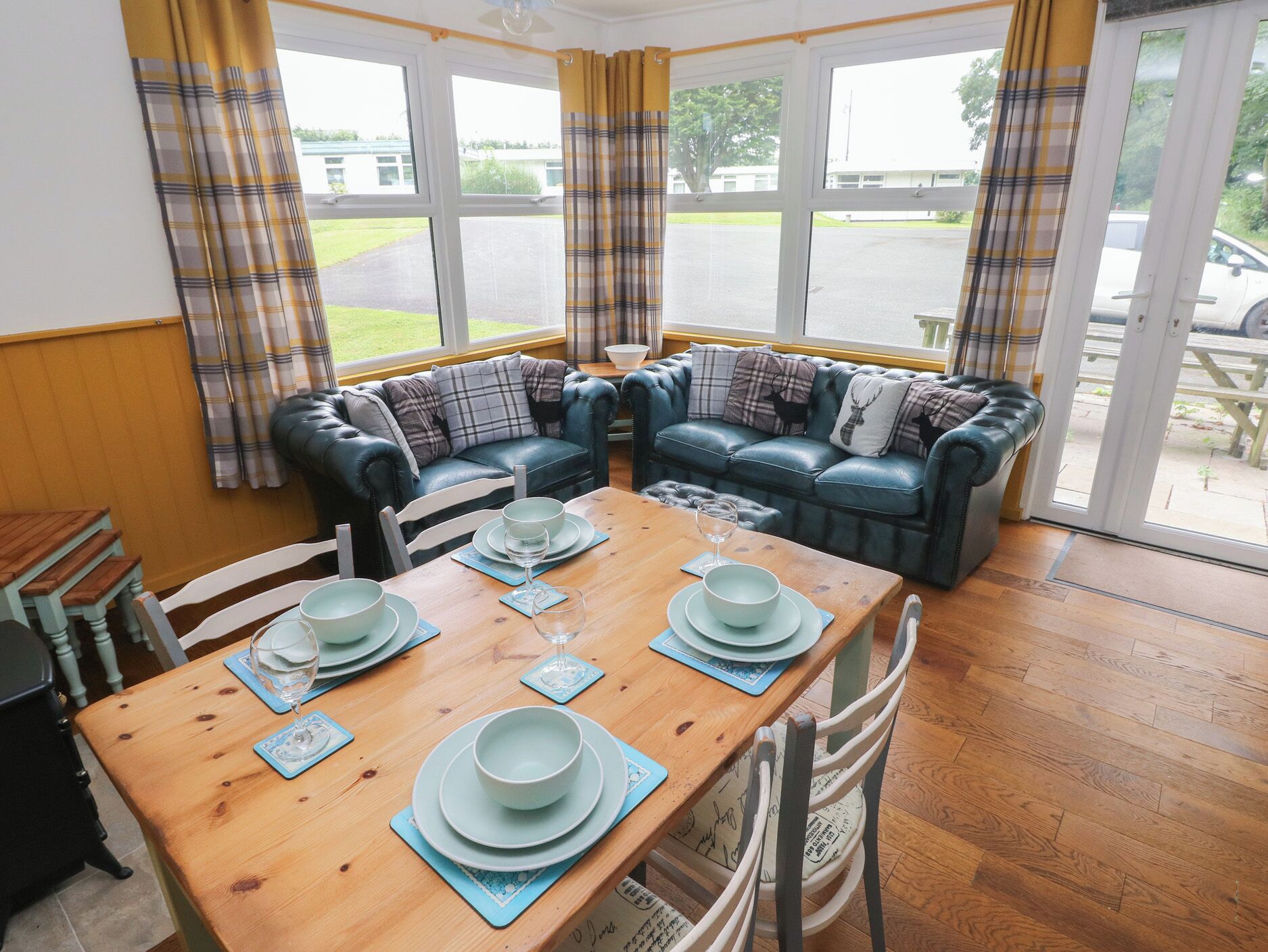 A cozy room with a wooden dining table set for four, a blue leather sofa, and a view of a parking lot through large windows.