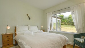 Ein Schlafzimmer mit einem großen Bett, Nachttischen aus Holz, einem Korbstuhl und einem Fenster mit Vorhängen.