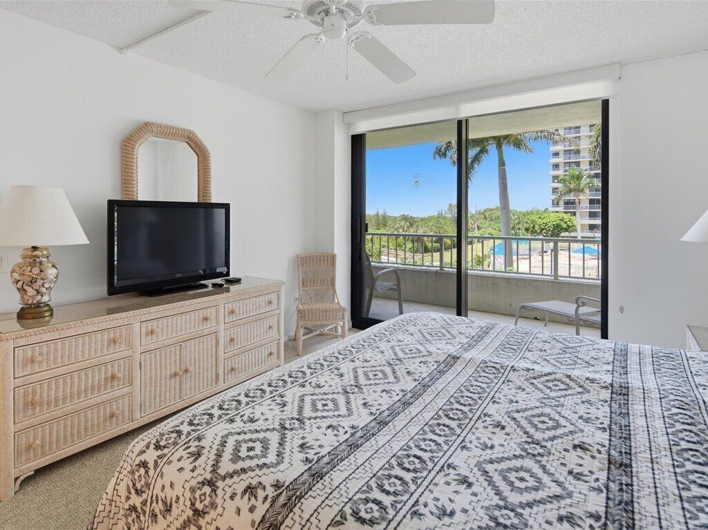 Habitación de hotel con cama, televisor en una cómoda, una silla y balcón con vistas a palmeras y una piscina.