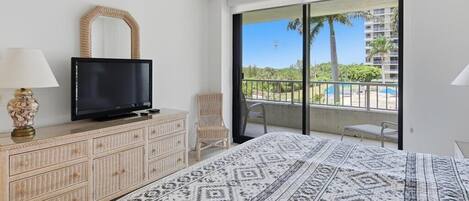 Habitación de hotel con cama, televisor en una cómoda, una silla y balcón con vistas a palmeras y una piscina.