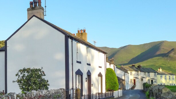 Une rue étroite bordée de bâtiments blancs, de murs de pierre et dominée par une chaîne de montagnes en arrière-plan.