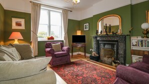 Un salon avec une cheminée, des murs verts, une grande fenêtre avec des rideaux, un canapé, un fauteuil et une bibliothèque.