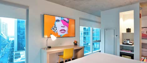 A modern hotel room with a large bed, a desk with a chair, a television mounted on the wall, and a large window offering a city view.