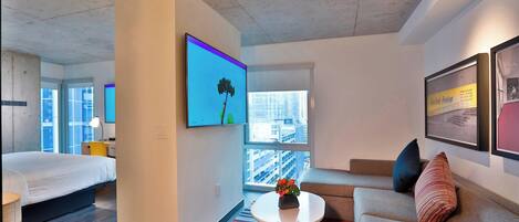 Una habitación de hotel moderna con una cama, un sofá, una mesita de centro con flores, un televisor y un ventanal con vistas a la ciudad.