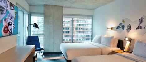 Une chambre d’hôtel avec deux lits, un bureau, une chaise, un tapis rayé aux couleurs vives et de grandes fenêtres donnant sur la ville.