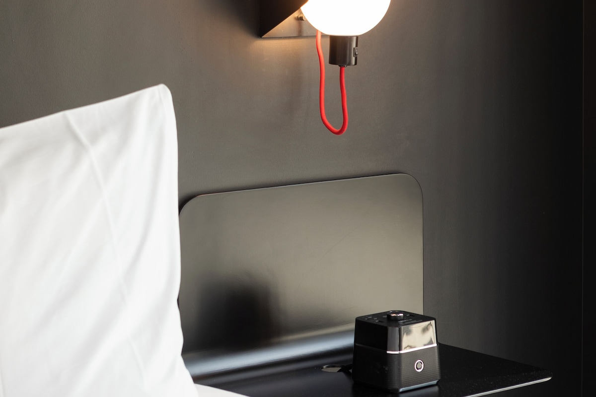 A modern hotel room with a neatly made bed, a bedside table with a camera, and a wall-mounted light fixture.