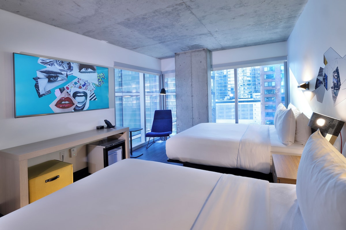 Une chambre d’hôtel moderne avec deux lits, un bureau, une chaise et de grandes fenêtres donnant sur la ville.